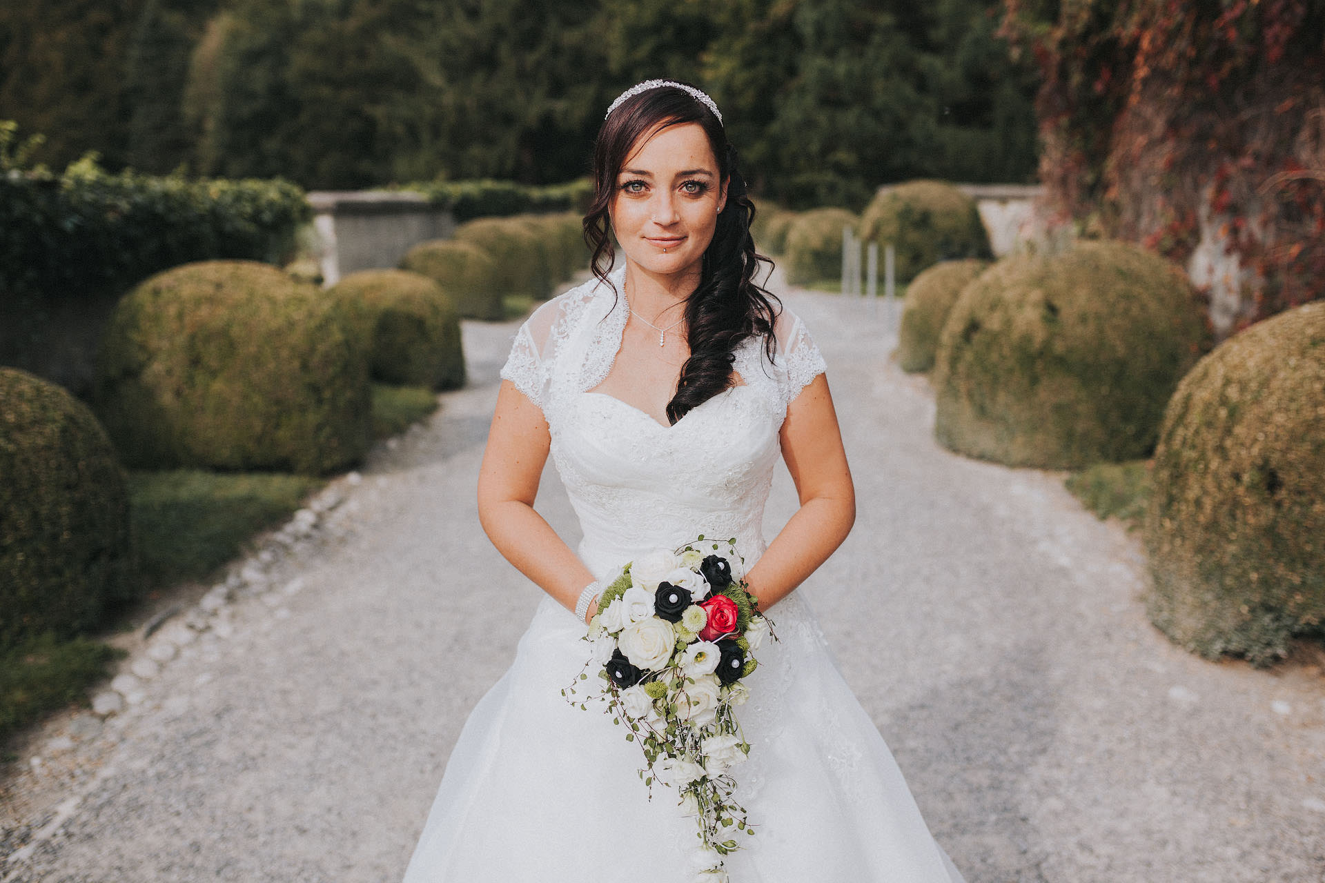 Braut hält einen Hochzeitsstrauß und blickt glücklich in die Kamera des Hochzeitsfotogafen bei Mittagssonne im Schlosspark des Schloss Ambras in Innsbruck in Tirol